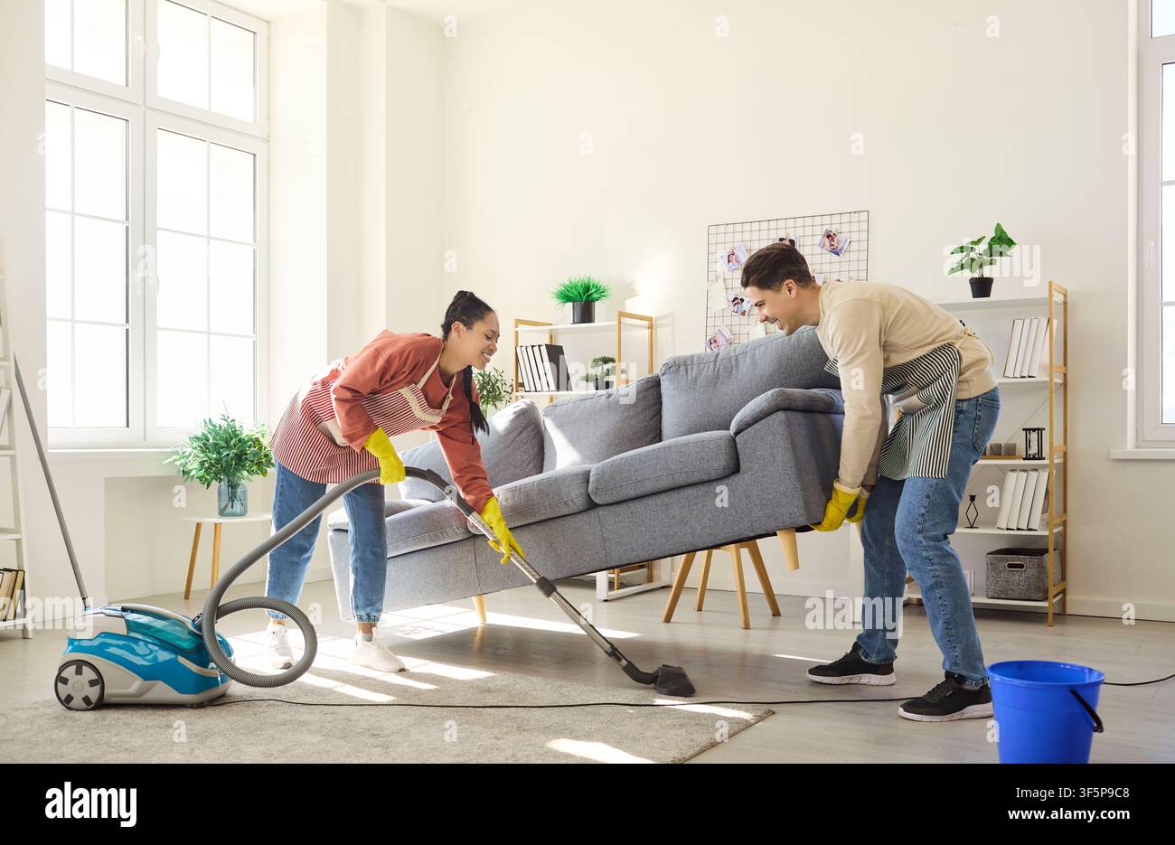 Couple vacuuming under sofa while lifting couch in bright modern living room Stock Photo