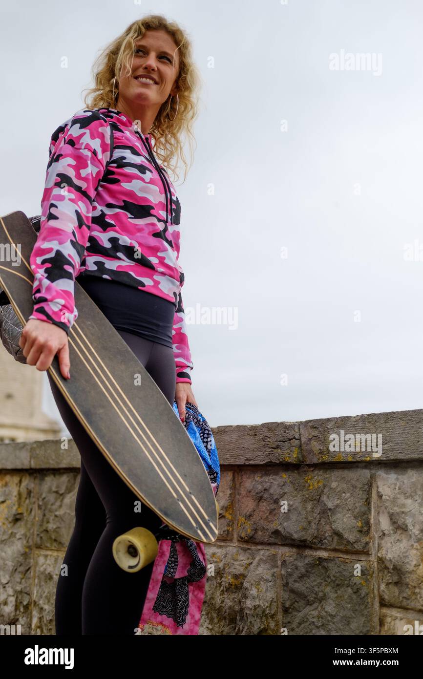 Young woman standing with longboard, smiling, dressed in pink camouflage hoodie Stock Photo