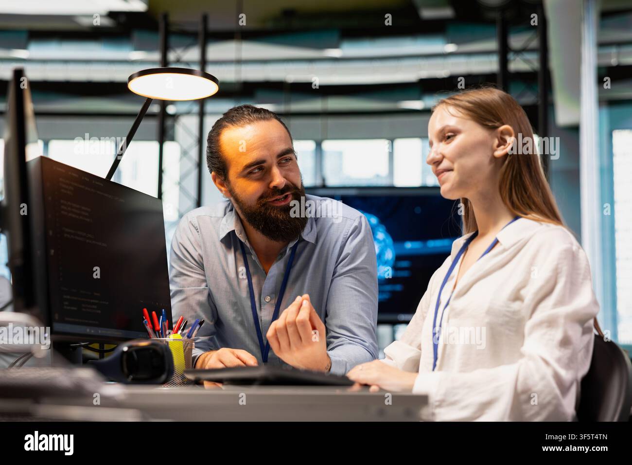 Friendly coworker having fun with young programmer while using artificial intelligence in office. Joyful colleague guiding junior dev through AI tool integration, mentoring her on using deep learning Stock Photo