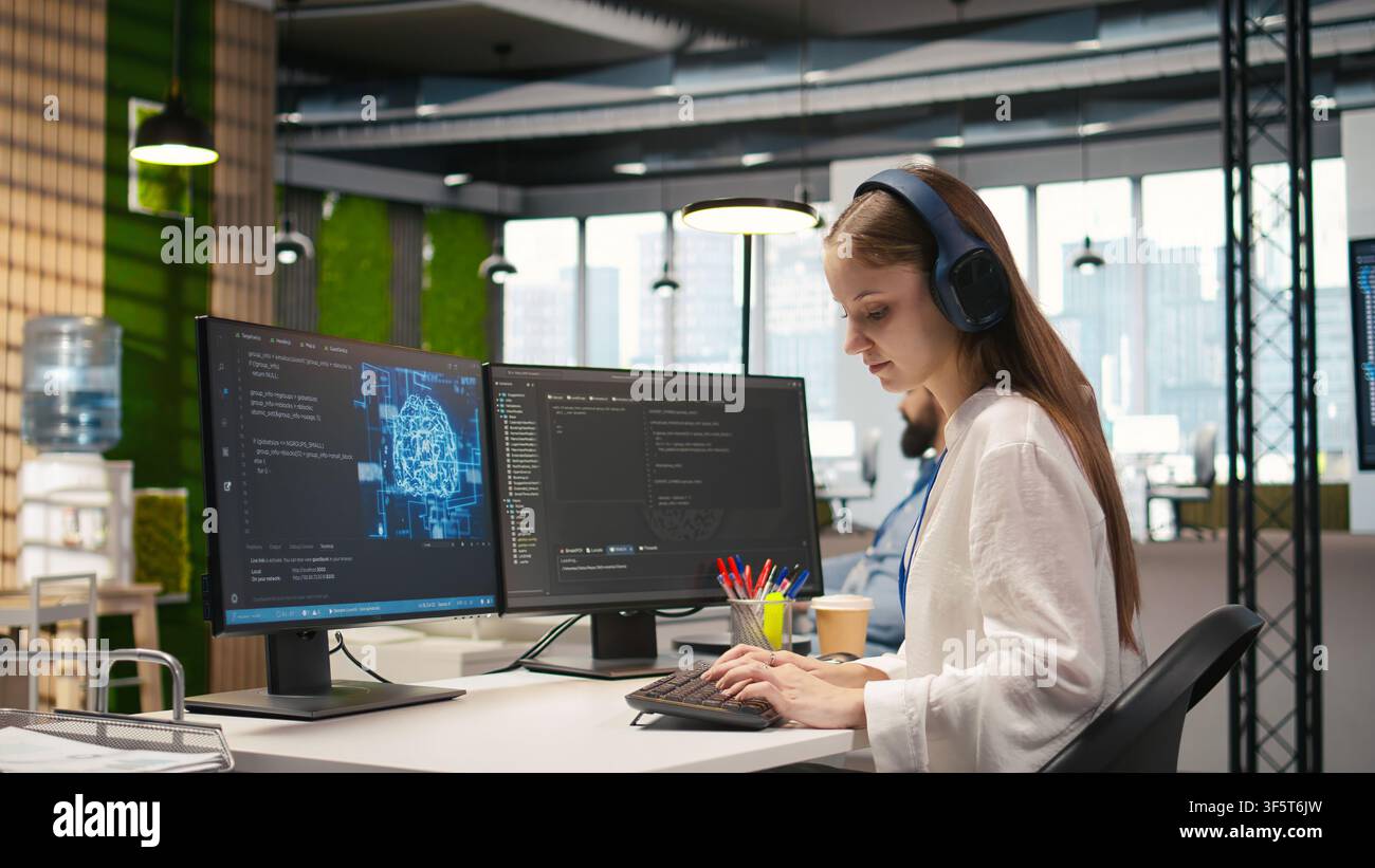 Junior dev developing software applications, optimizing code for performance, listening music. Young female engineer wearing headphones in office using AI to resolve bottlenecks, camera A Stock Photo
