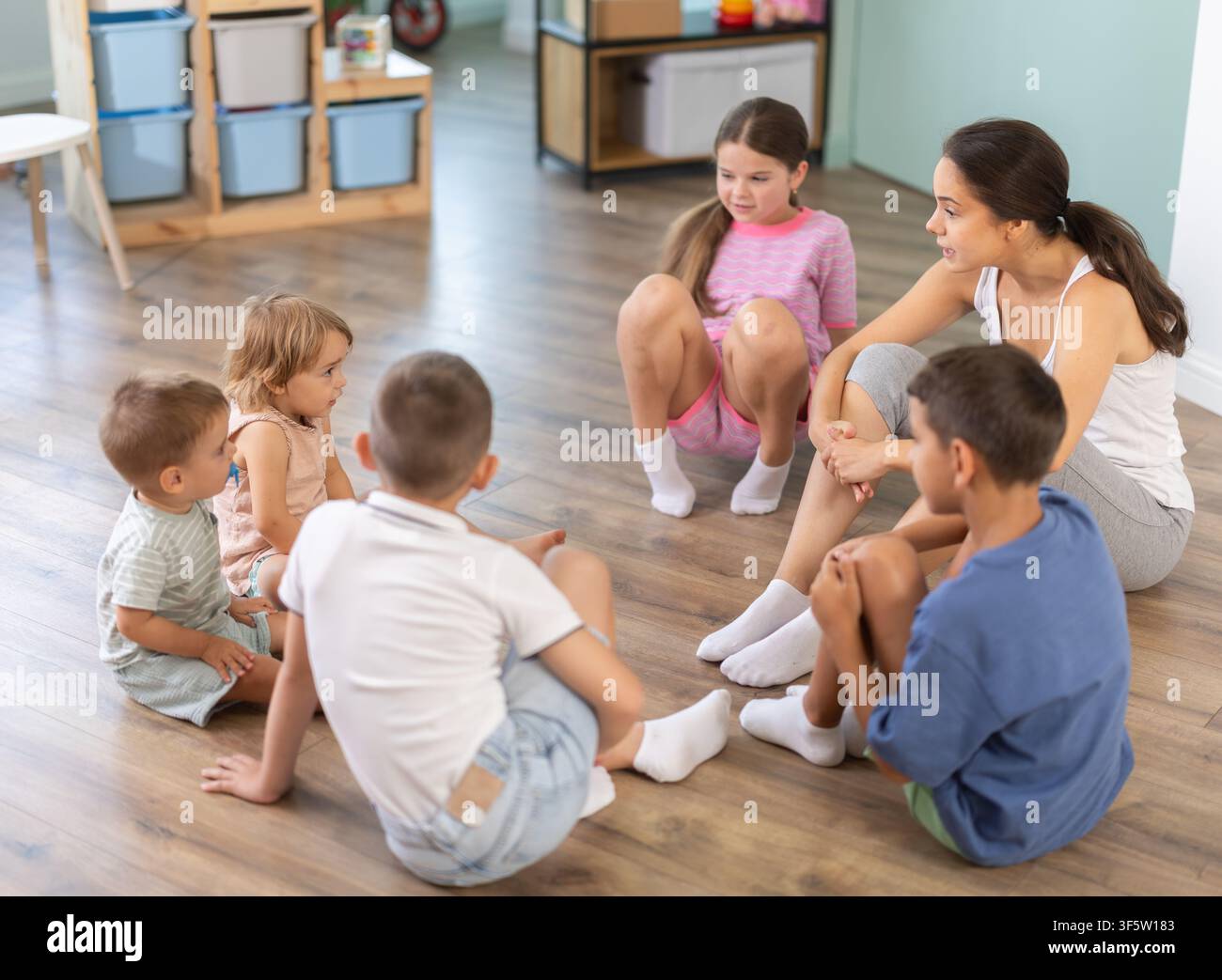Children sitting on floor while caring female teacher explains lesson in kindergarten Stock Photo