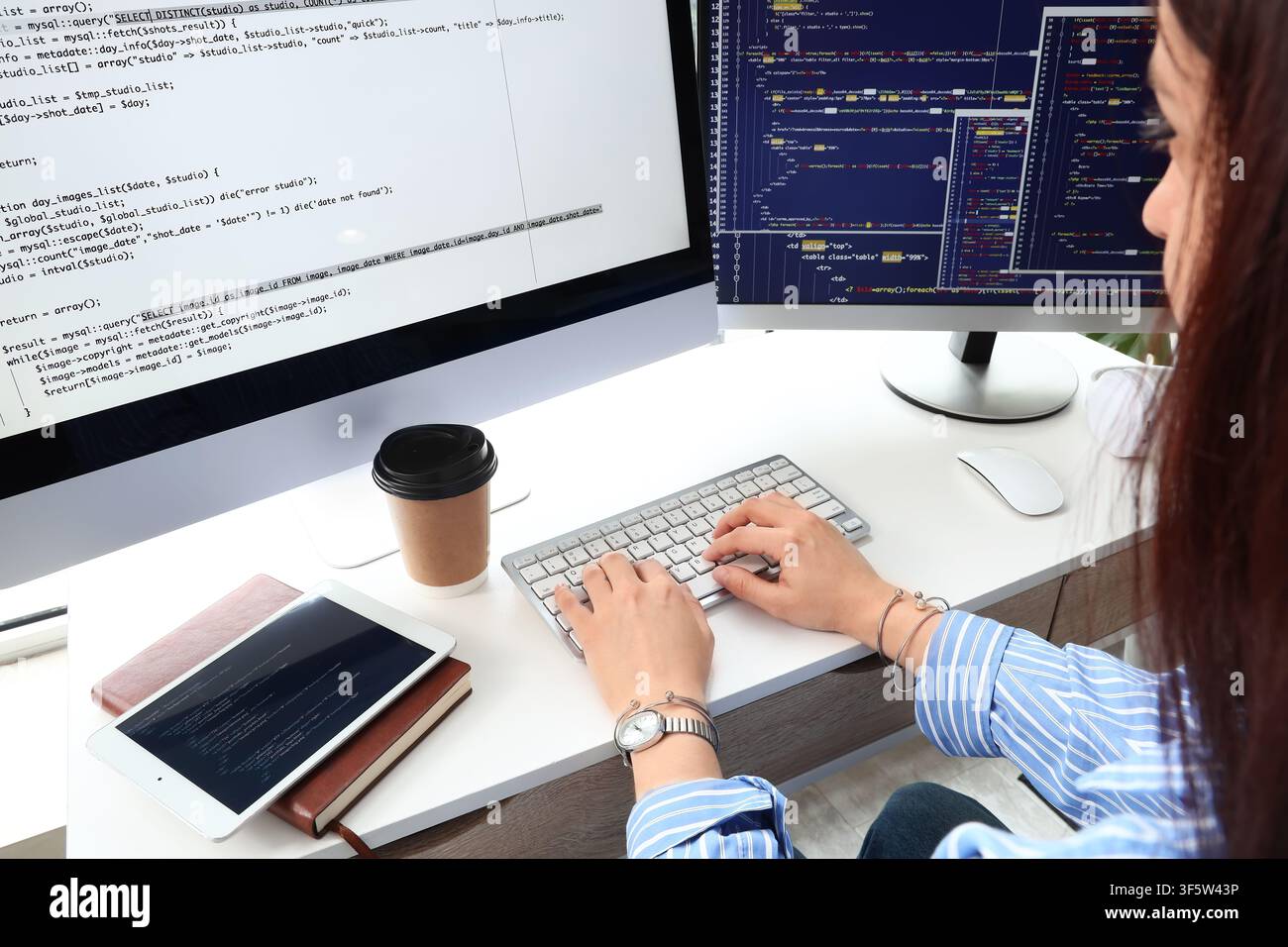 Female programmer coding on a desktop computer with multiple screens displaying complex software development code, the womans hands are actively typin Stock Photo