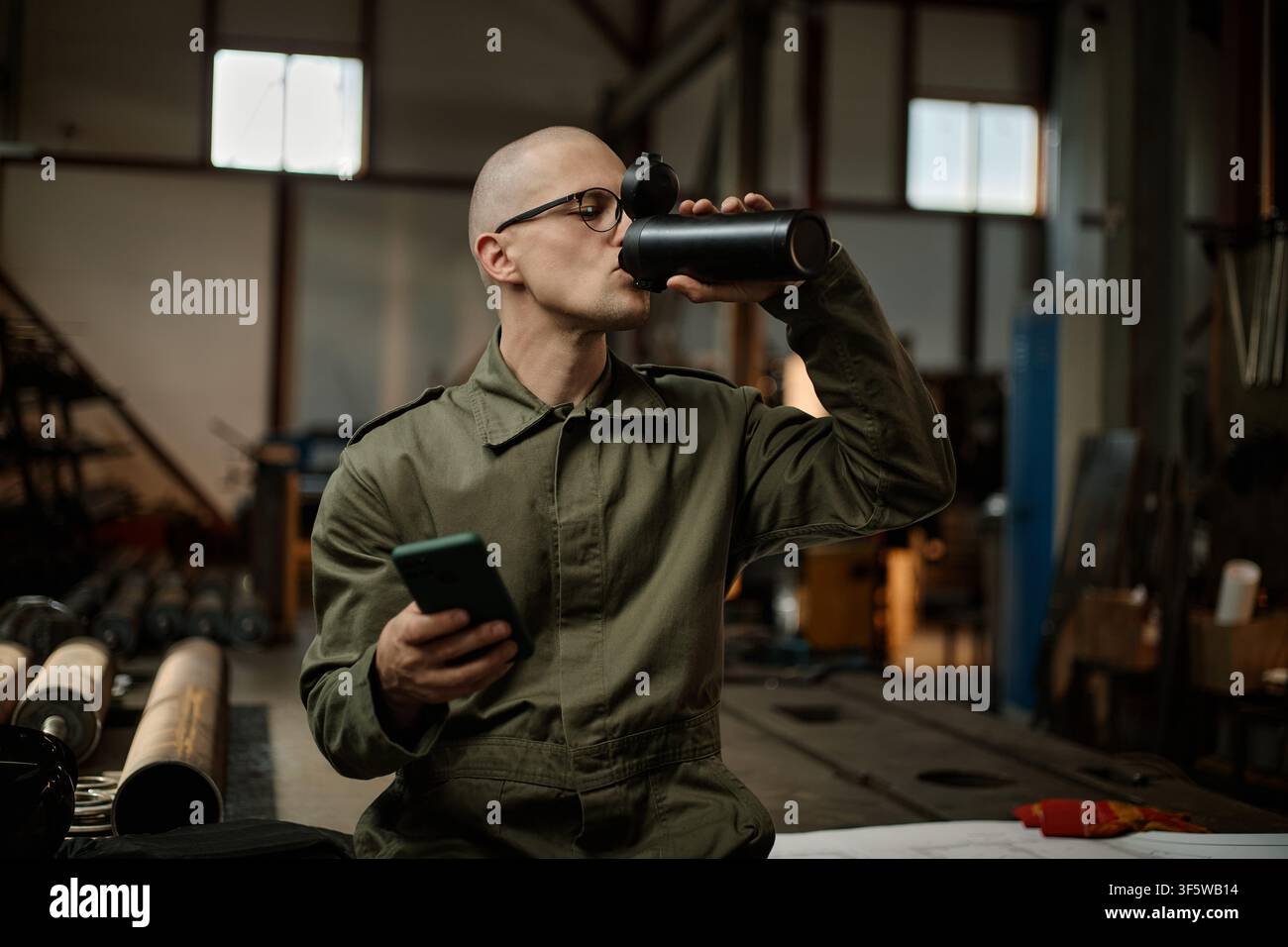 Male Welder Drinking Water and Using Smartphone during Work Break Stock Photo