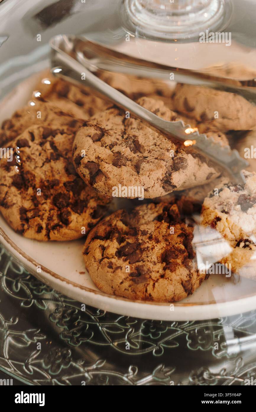 Fresh chocolate chip cookies displayed under a glass dome with serving tongs in a modern cafe. Homemade pastries presented for customers. Stock Photo