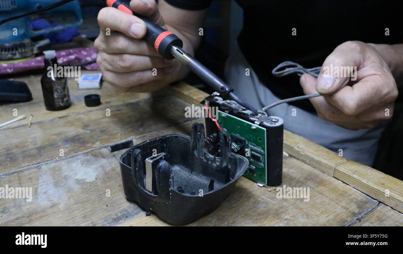 mechanic repairs a battery pack in a workshop. He holds a soldering iron, carefully soldering contacts to the electronic circuit board with wires Stock Photo