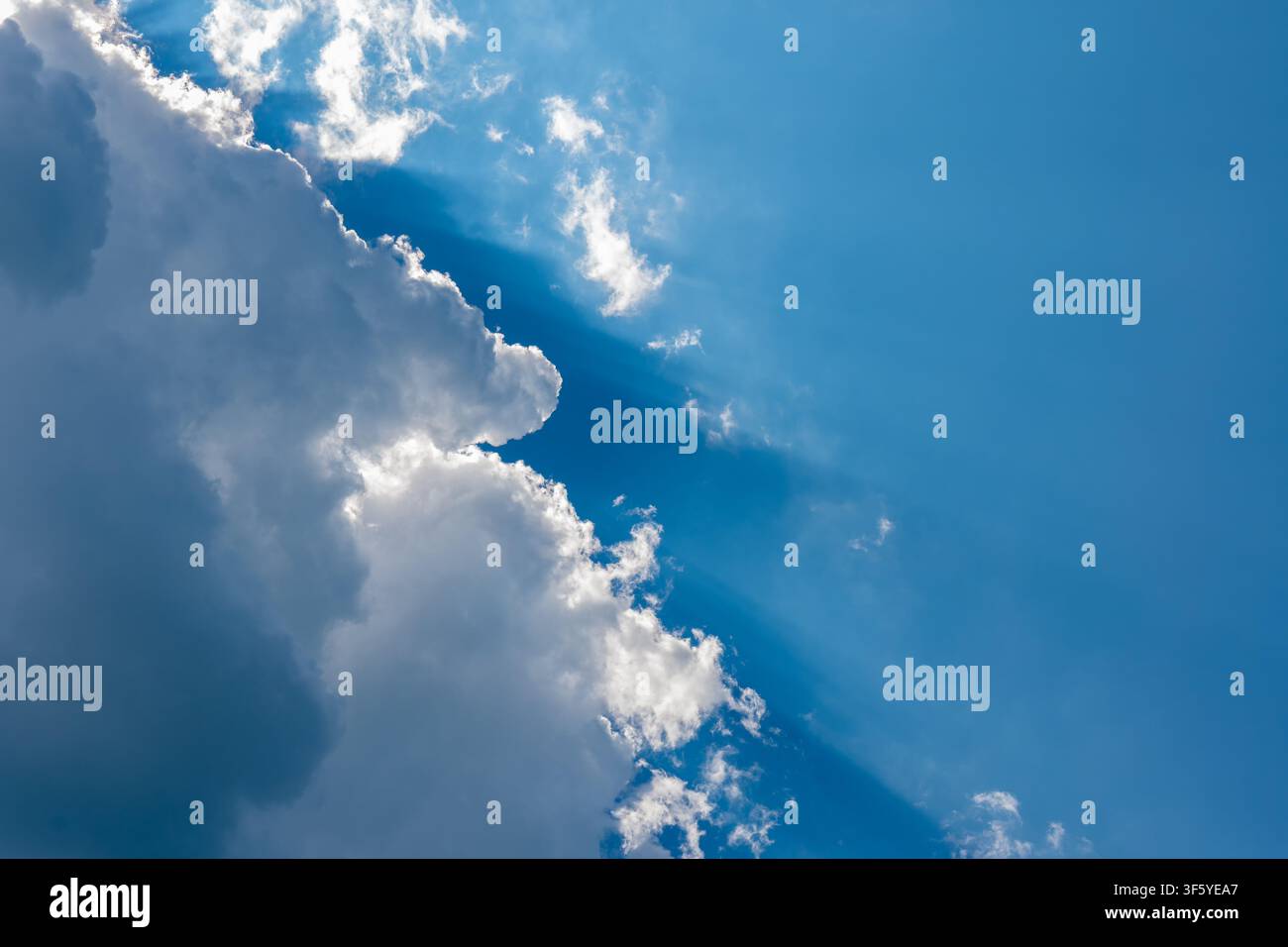 Bright sun rays stream through large white clouds in a clear blue sky. Stock Photo