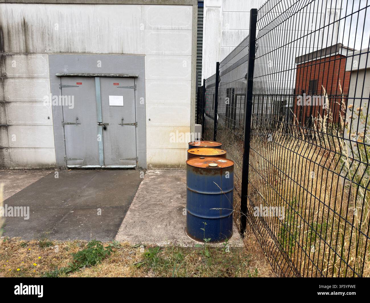 Three weathered industrial steel drums beside security fencing and warehouse loading doors in an anonymous British industrial estate. Stock Photo