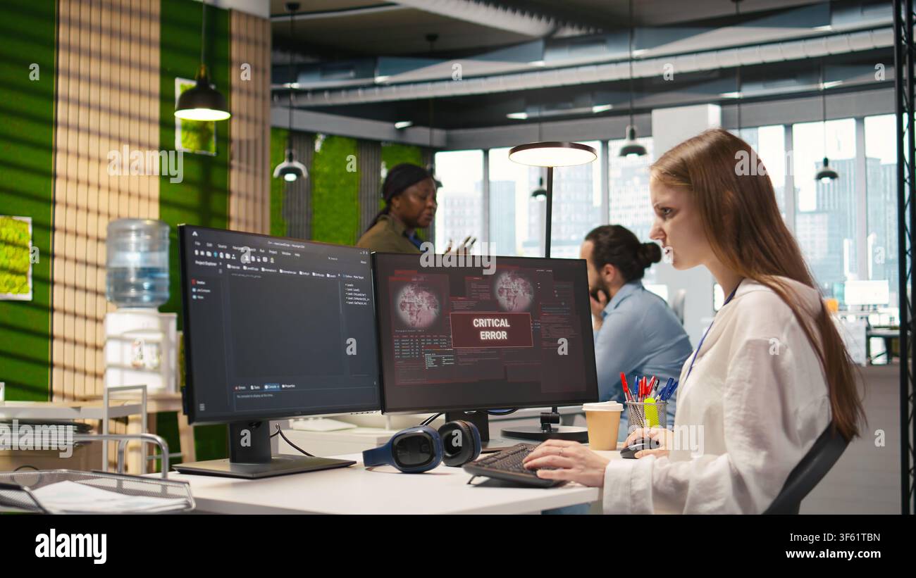 Female programmer in startup office struggling to fix application errors while coding. Young woman dealing with computer bugs while coding in software development agency, camera A Stock Photo