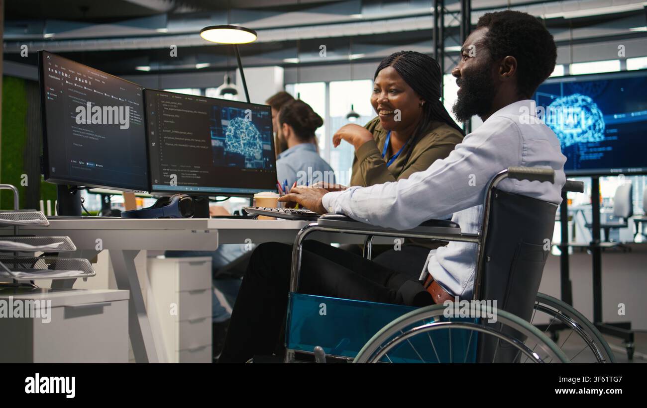 Joyous technician with disability and coworker chatting while coding AI algorithms debugging software issues. Upbeat tech startup office IT expert in wheelchair and colleague using computer, camera A Stock Photo