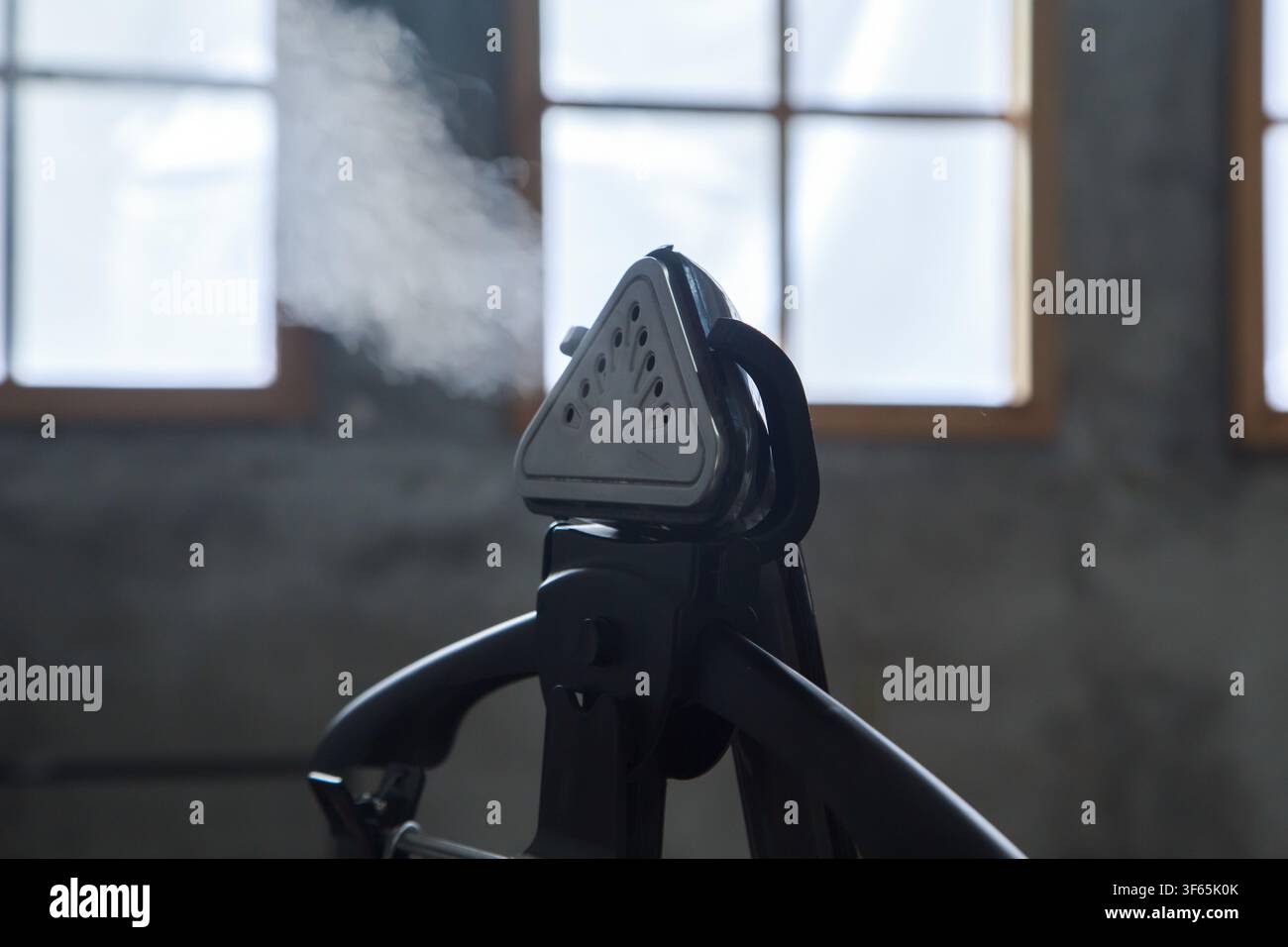 Handheld garment steamer emitting a plume of white steam on a black clothes hanger in a modern industrial setting. Stock Photo