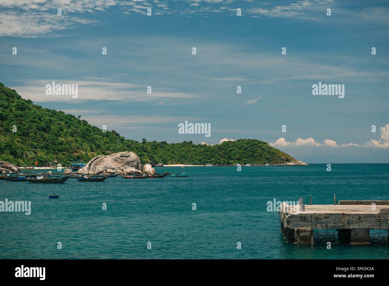 A concrete dock stretches into calm turquoise ocean water near a green hillside shoreline dotted with small anchored vessels under a blue sky. Stock Photo