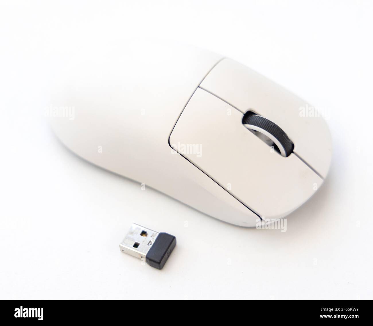A white wireless computer mouse is placed on a white surface alongside a small black and silver USB receiver Stock Photo