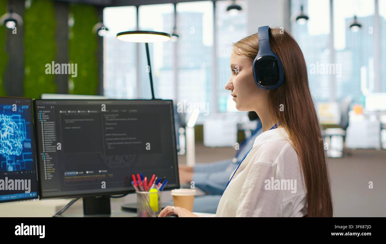 Junior dev developing software applications, optimizing code for performance, listening music. Young female engineer wearing headphones in office using AI to resolve bottlenecks, camera A Stock Photo