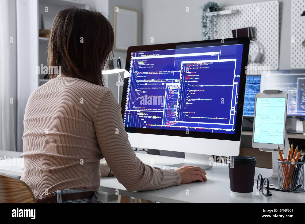 Female software developer coding on a desktop computer with multiple screens in a modern home office setting Stock Photo