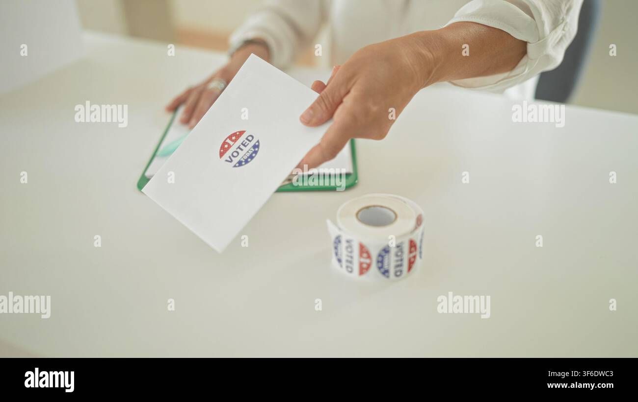 Woman hand offering ballot envelope beside roll of voted stickers and clipboard on table at building polling station; pride civic duty. Stock Photo