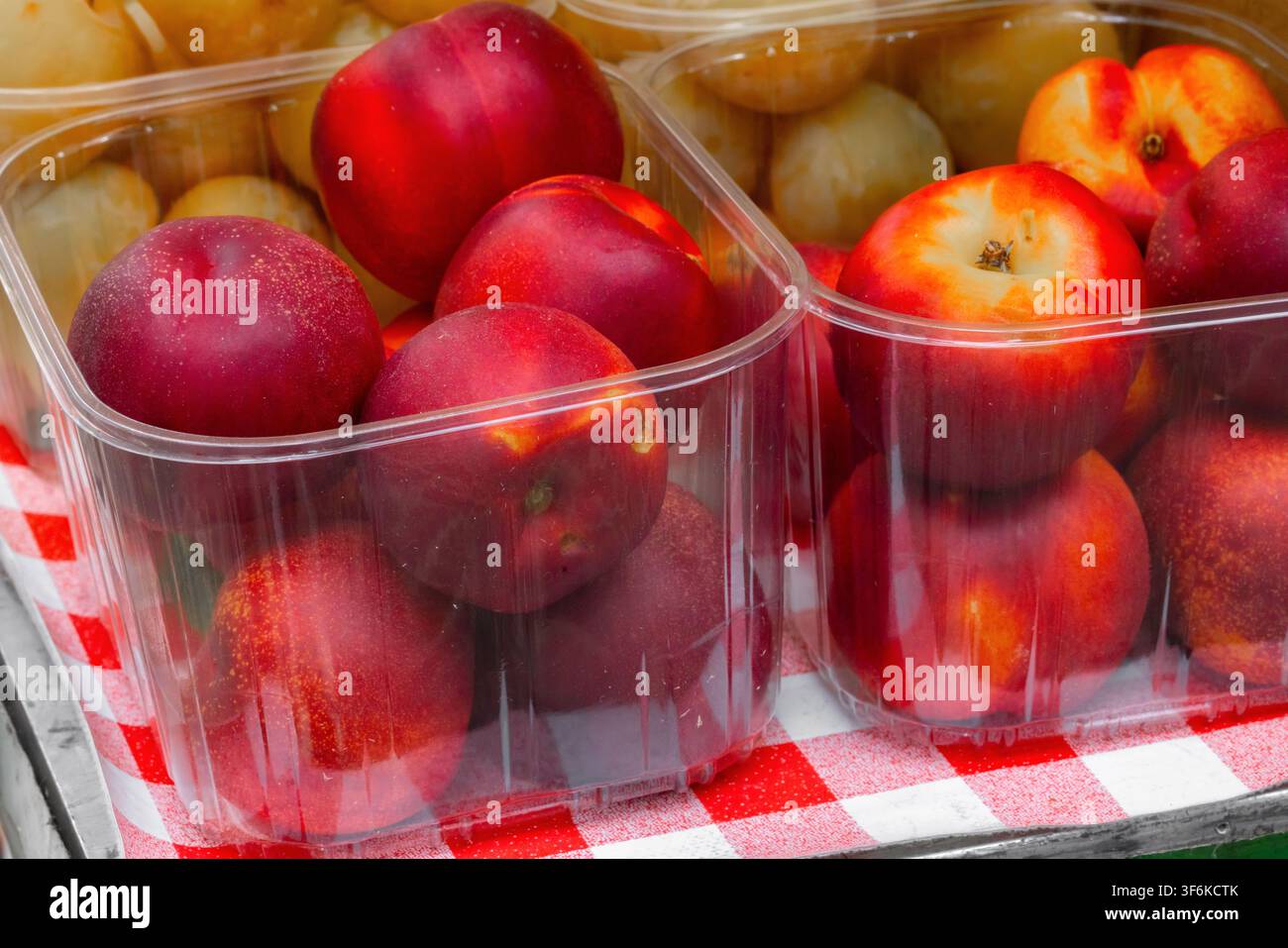 Red nectarines and yellow plums are arranged in transparent containers on a checkered tablecloth, showcasing fresh produce at a market setting Stock Photo