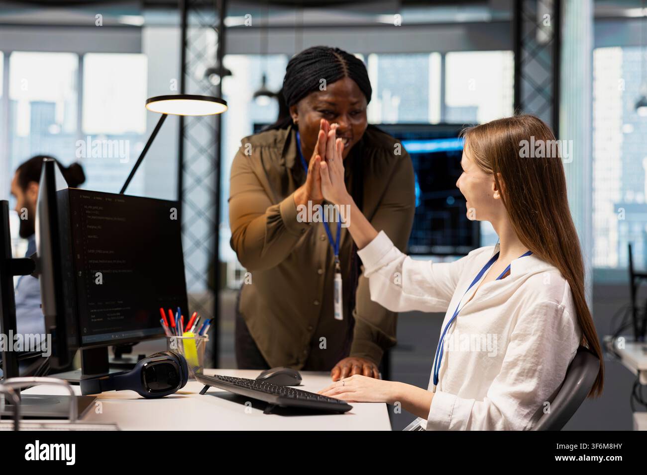 Smiling system administrator mentoring junior dev at desk in office, congratulating her for optimizing code. Joyous tech startup IT specialist doing high five with female developer intern Stock Photo