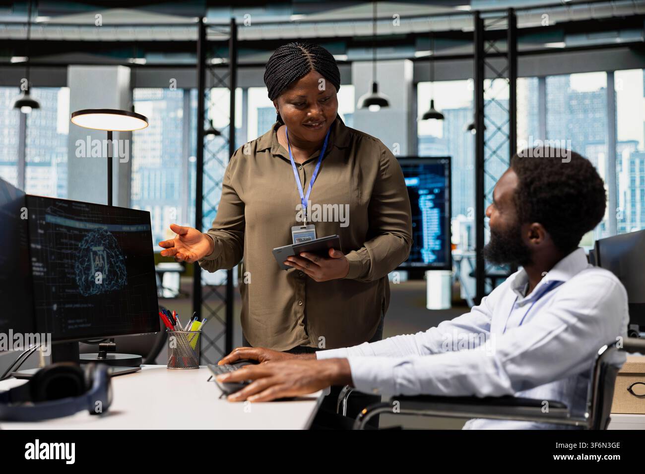 Smiling manager oversees software developer in wheelchair using AI automation, refining algorithms. Jolly supervisor pleased with work done by programmer with paraplegia using artificial intelligence Stock Photo