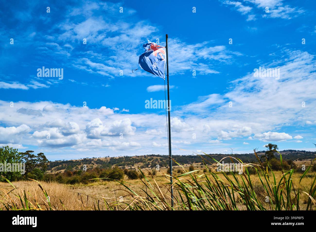 A worn, weathered, iconic, old Australian flag flying in a rural, country field against blue sky and clouds. In Collector, NSW, Australia. Stock Photo