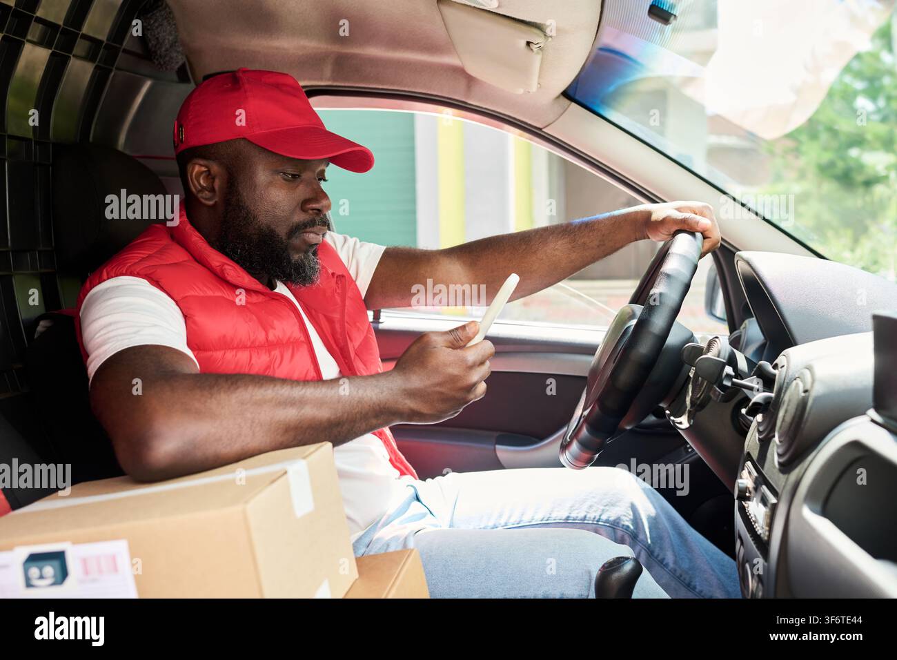 Black Courier Man Using Smartphone App While Driving Delivery Van Stock Photo