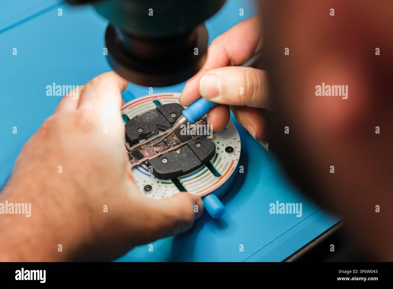 Technician repairs electronic circuit board using precision tools on a blue work surface with a microscope in the background Stock Photo