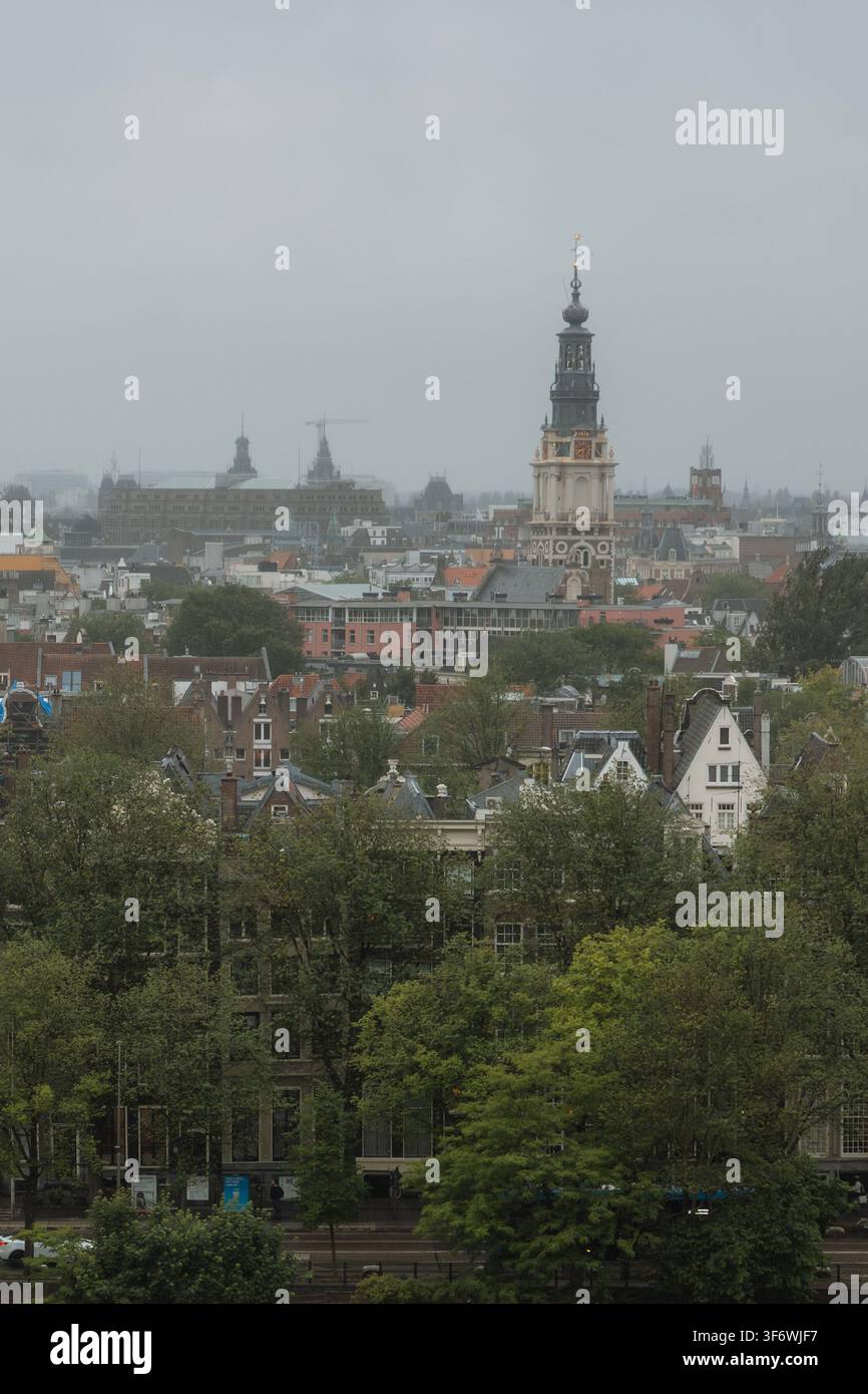 Amsterdam skyline capturing the Zuiderkerk bell tower rising above traditional Dutch canal houses and green trees under a cloudy sky Stock Photo