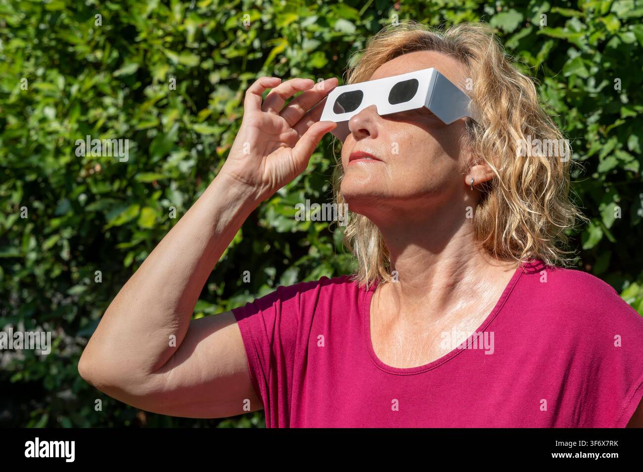 Blonde woman in red observes the sun with classic solar eclipse protective glasses Stock Photo
