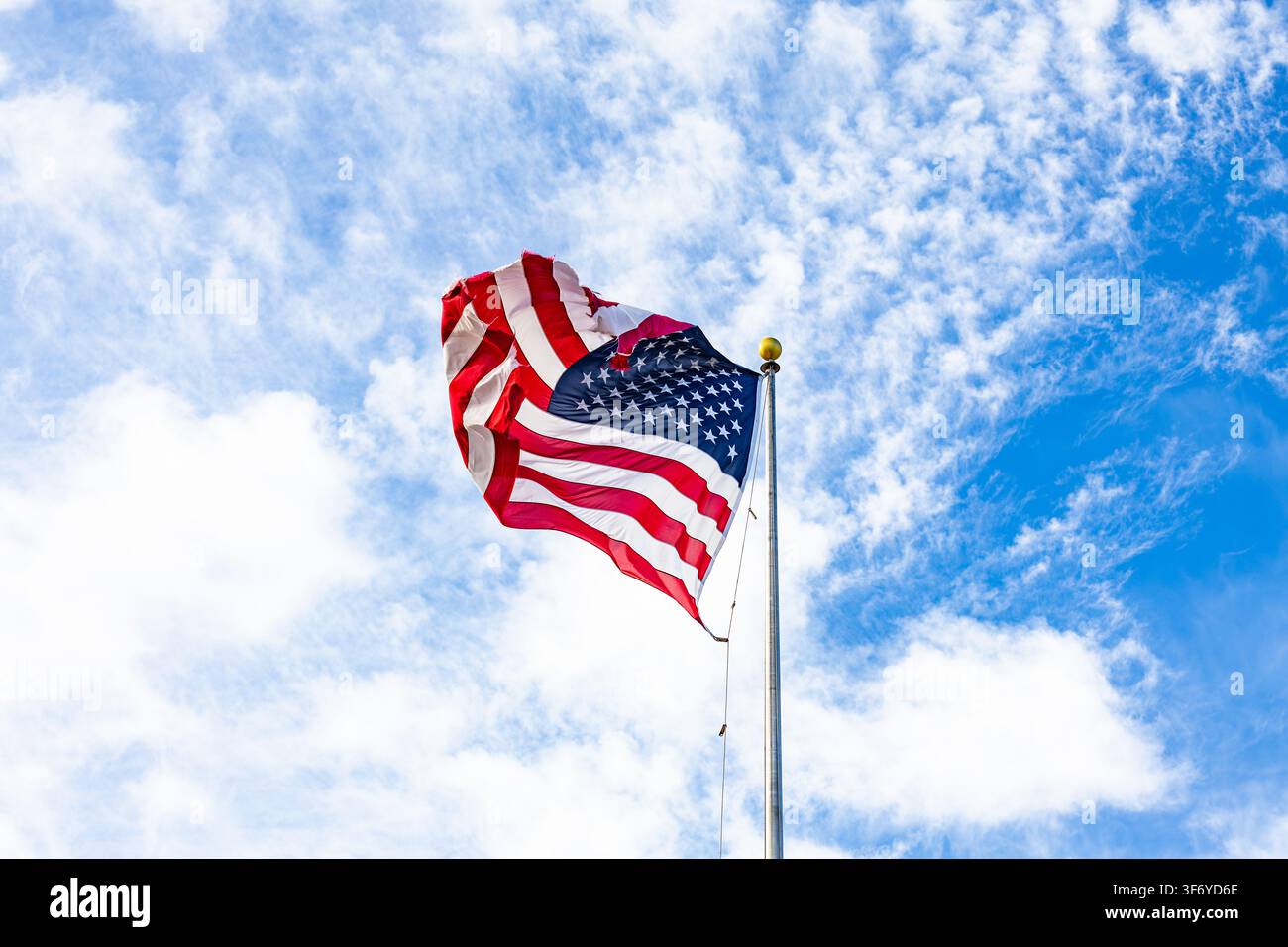 United States national flag waving on a metal flagpole against a bright blue sky with textured white clouds in broad daylight Stock Photo