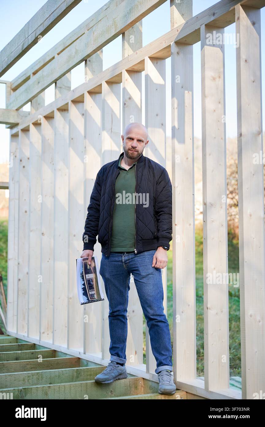 Builder stands inside timber wall frame holding project plans, overseeing construction progress and visualizing future layout of residential building Stock Photo