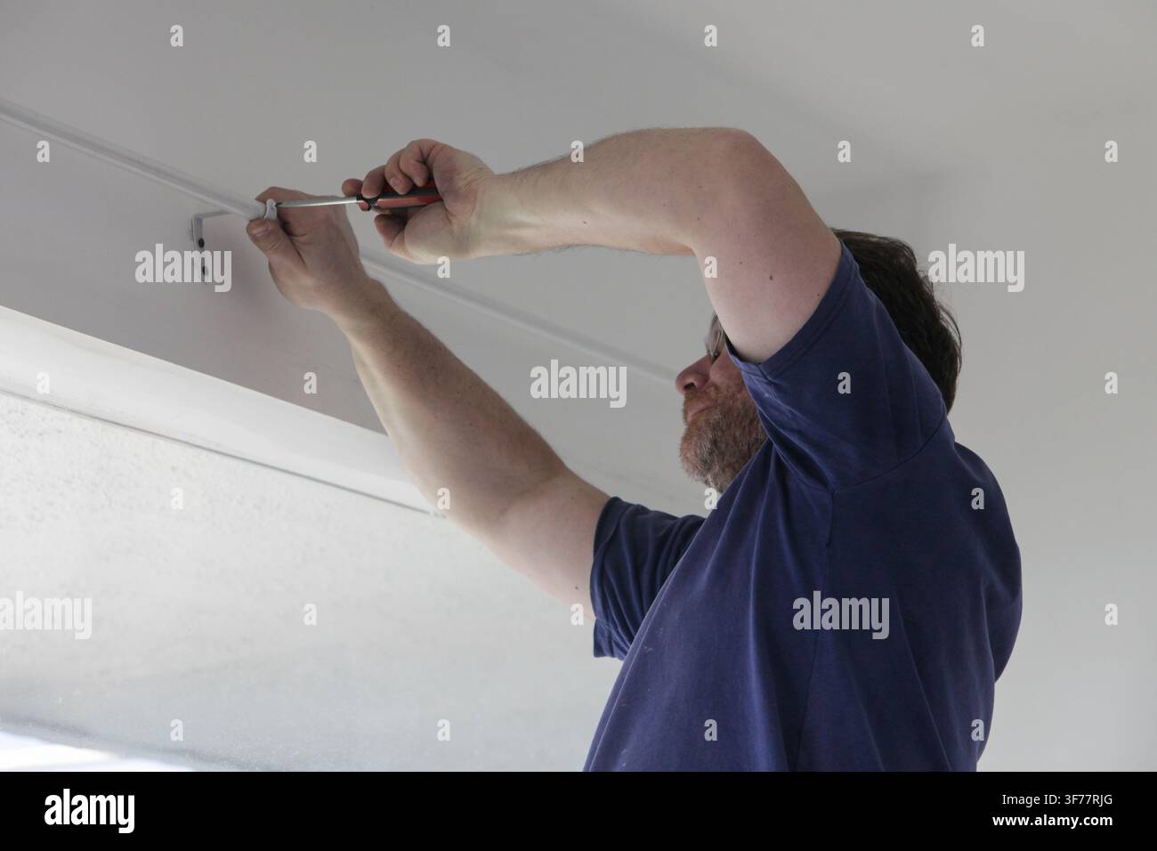 Man installing curtain rod support brackets with a screwdriver above a window during home improvement work. Practical DIY installation, home repair, r Stock Photo