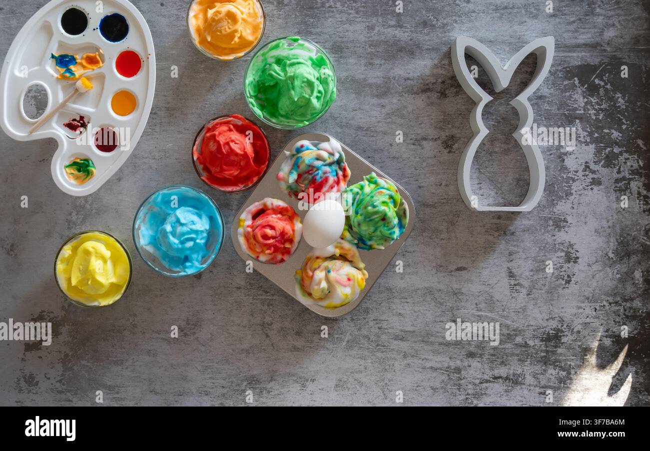 Overhead Easter craft composition featuring colorful painted eggs a watercolor palette and a white bunny decoration arranged on a textured gray backgr Stock Photo