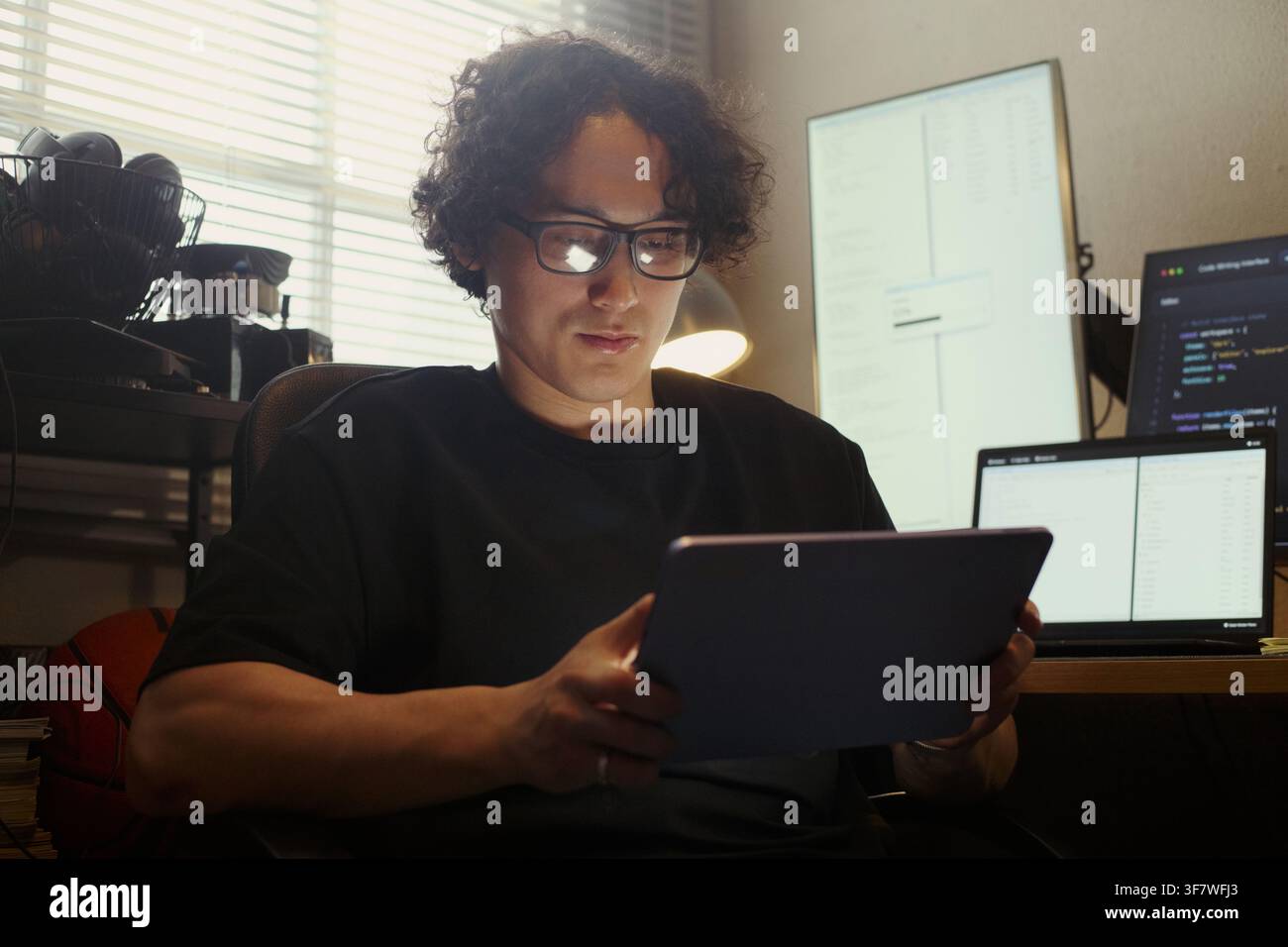 Asian young man wearing glasses using digital tablet in home office, coding on multiple screens in background, representing software development and remote tech work, medium shot Stock Photo