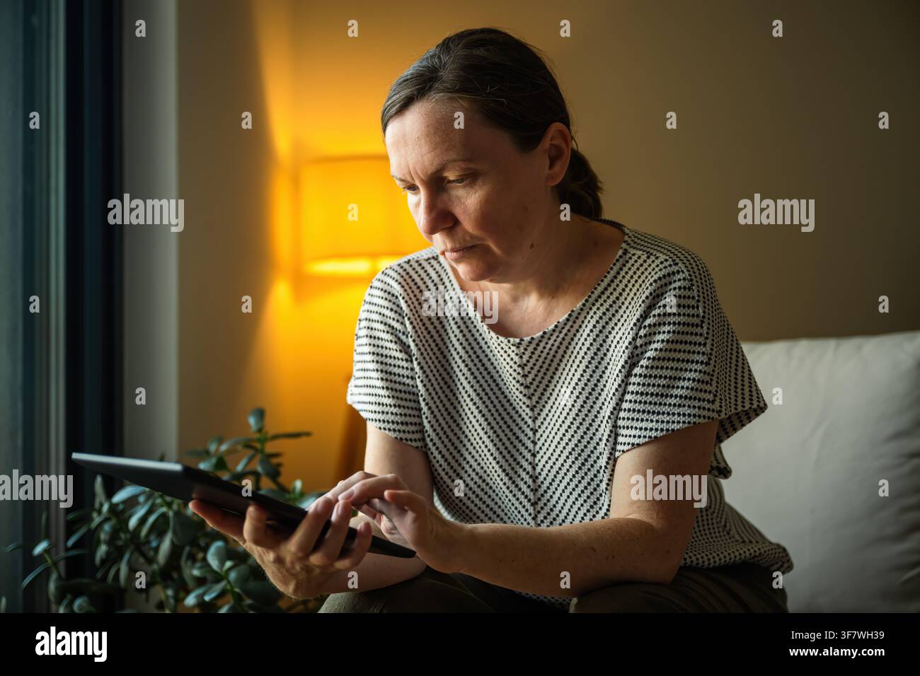 Woman using a tablet computer in a comfortable home setting during the evening. Digital lifestyle, remote work and online communication concept. Selec Stock Photo