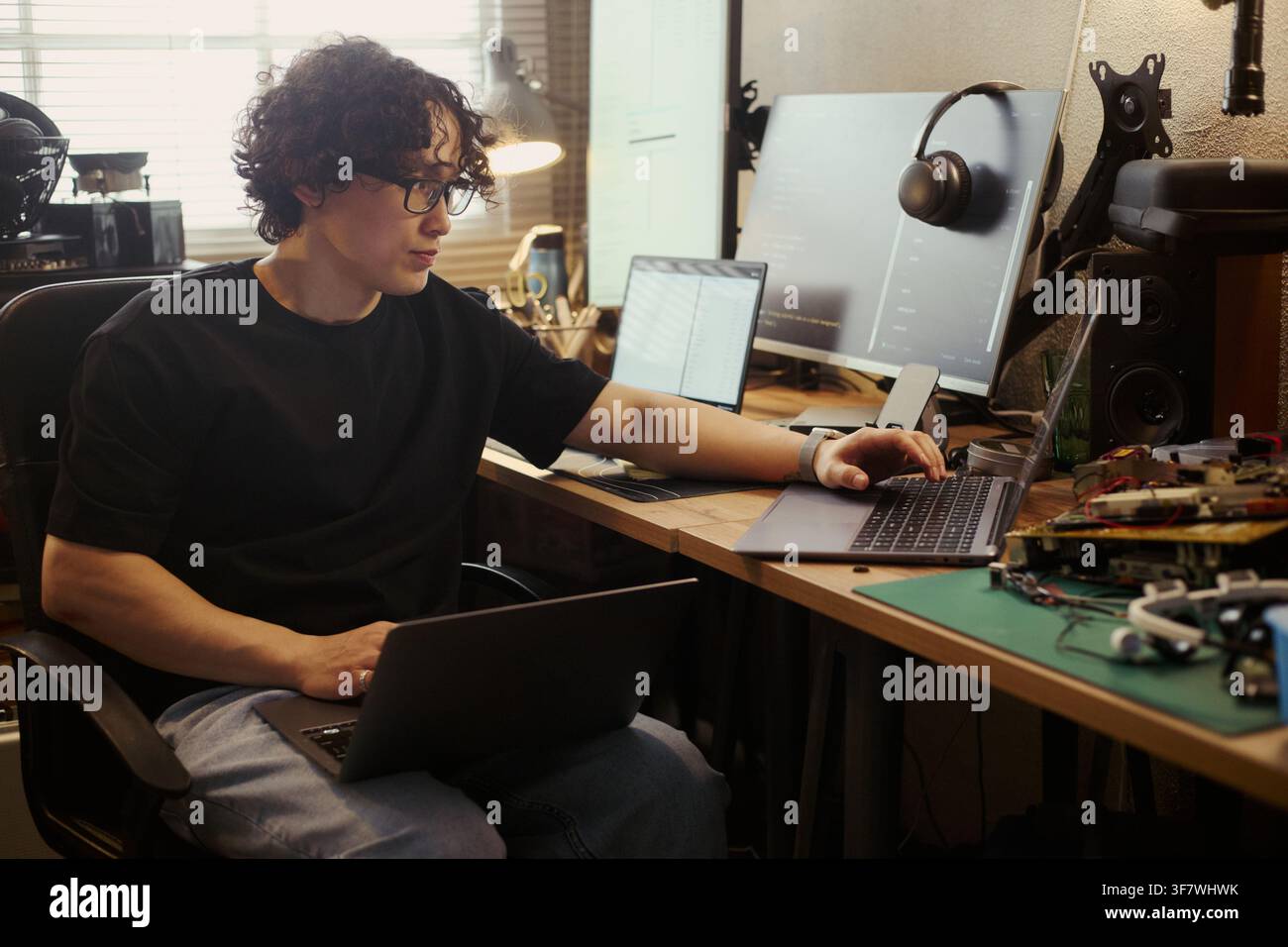 Asian young man working on multiple laptops at home office desk, coding and testing hardware. Image suitable for remote work, software development, and IT education campaigns Stock Photo