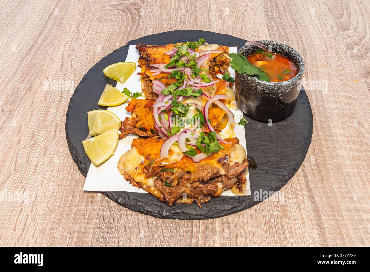 Close-up of birria-style quesatacos with cilantro, onion, lime wedges, and consommé on a slate plate. Stock Photo