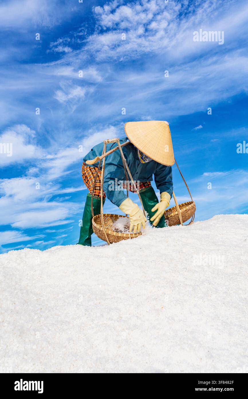 Worker in conical hat collects salt from a heap under a blue sky in Vietnam Stock Photo