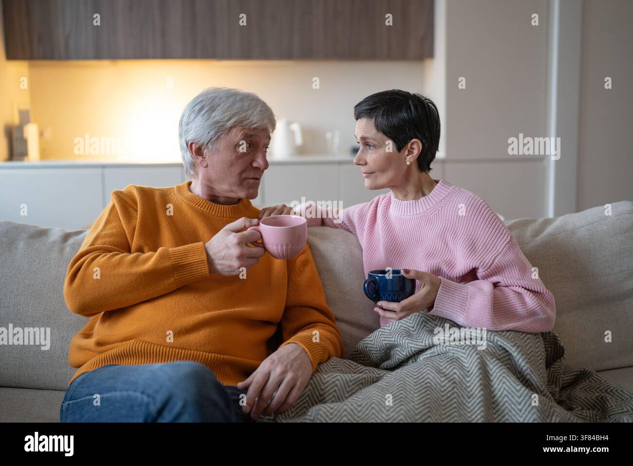 Senior husband and wife sitting on couch at home together drinking tea and talking with each other. Stock Photo