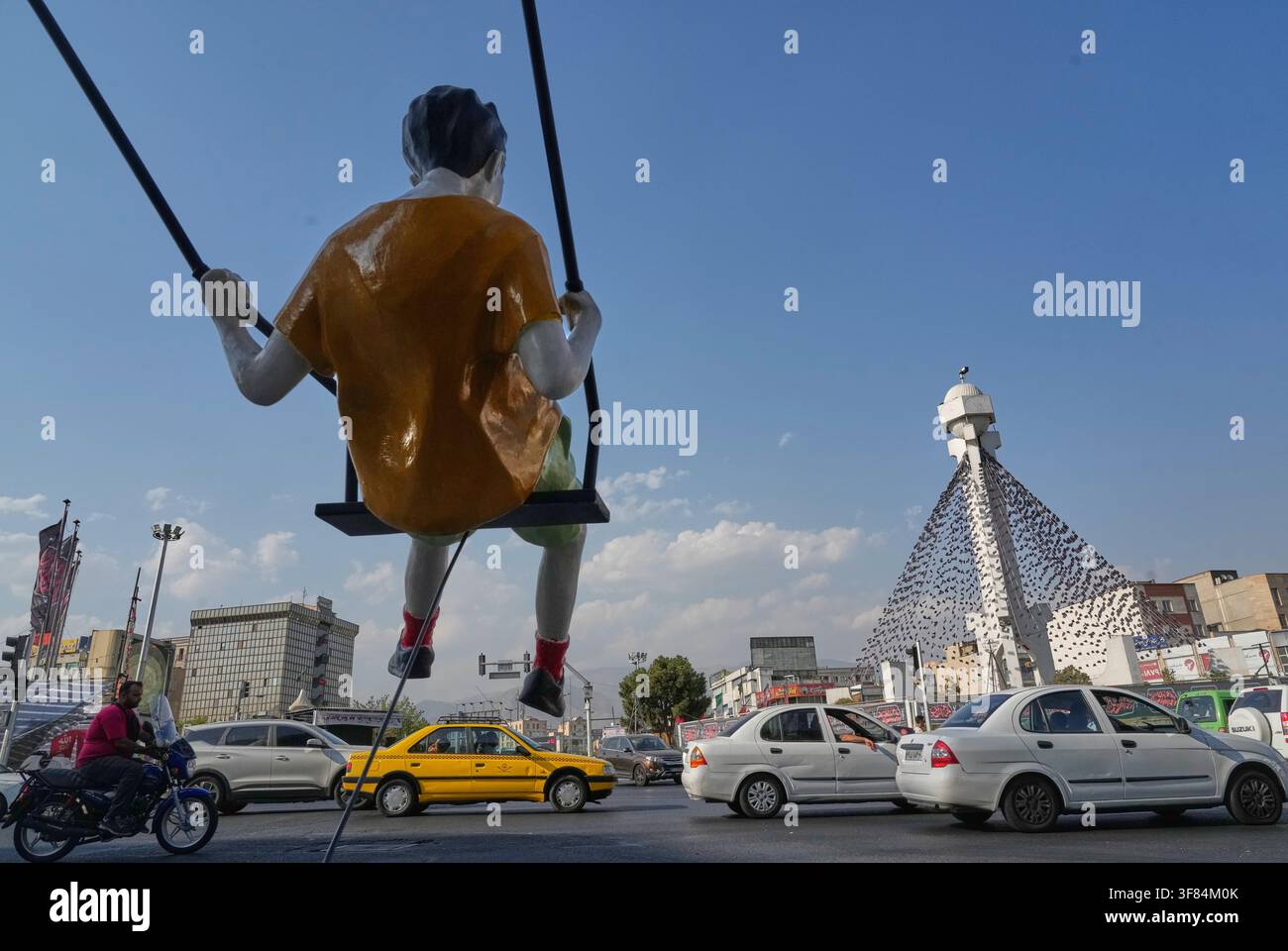 Vehicles drive past a statue of a swinging boy at a square in Tehran ...