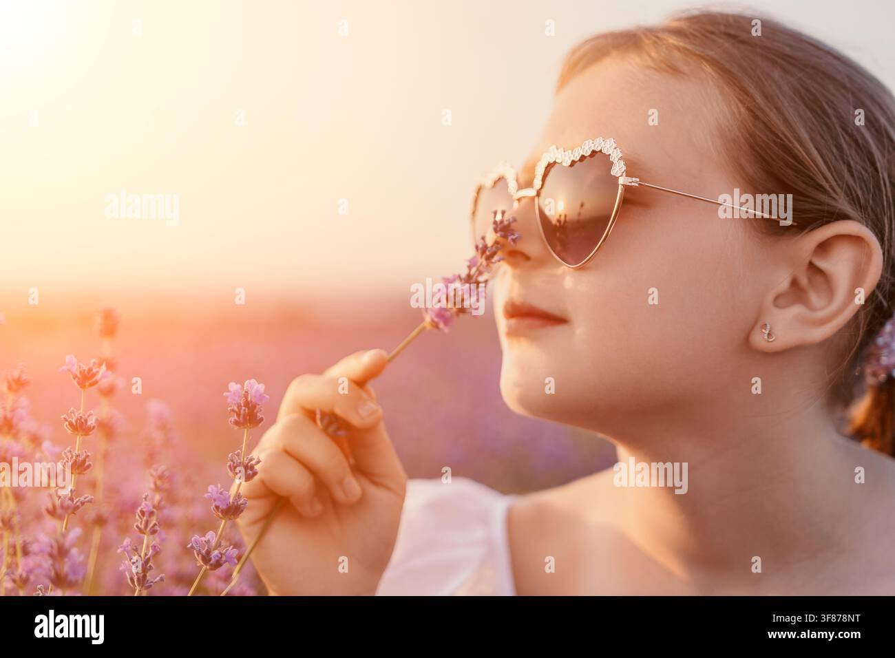 A young girl with heart-shaped sunglasses gently smells a sprig of ...