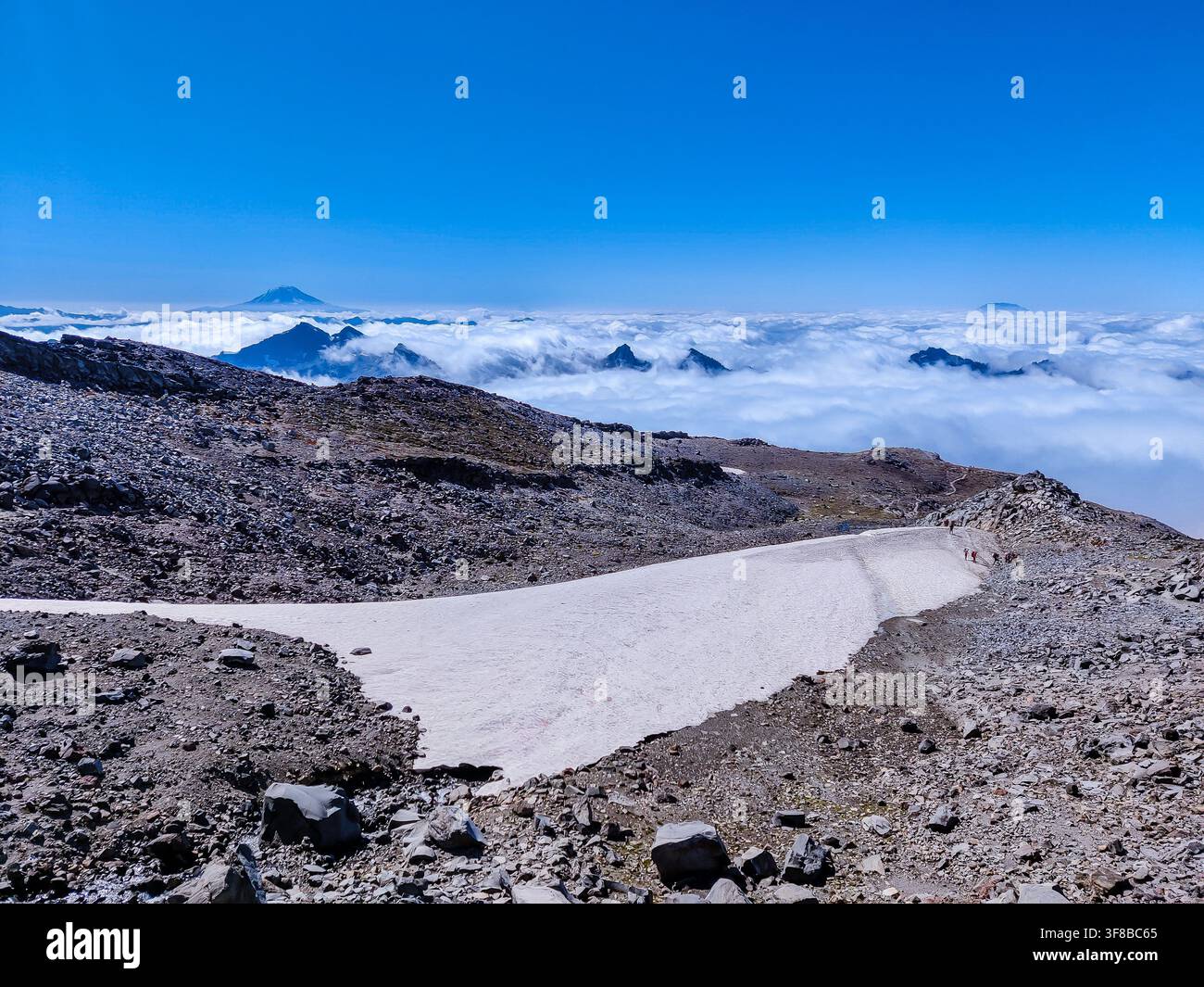 Clear blue sky above rugged mountain peak. Snow patch lines rocky terrain, untouched and pristine. Clouds blanket valley below like a soft white sea Stock Photo