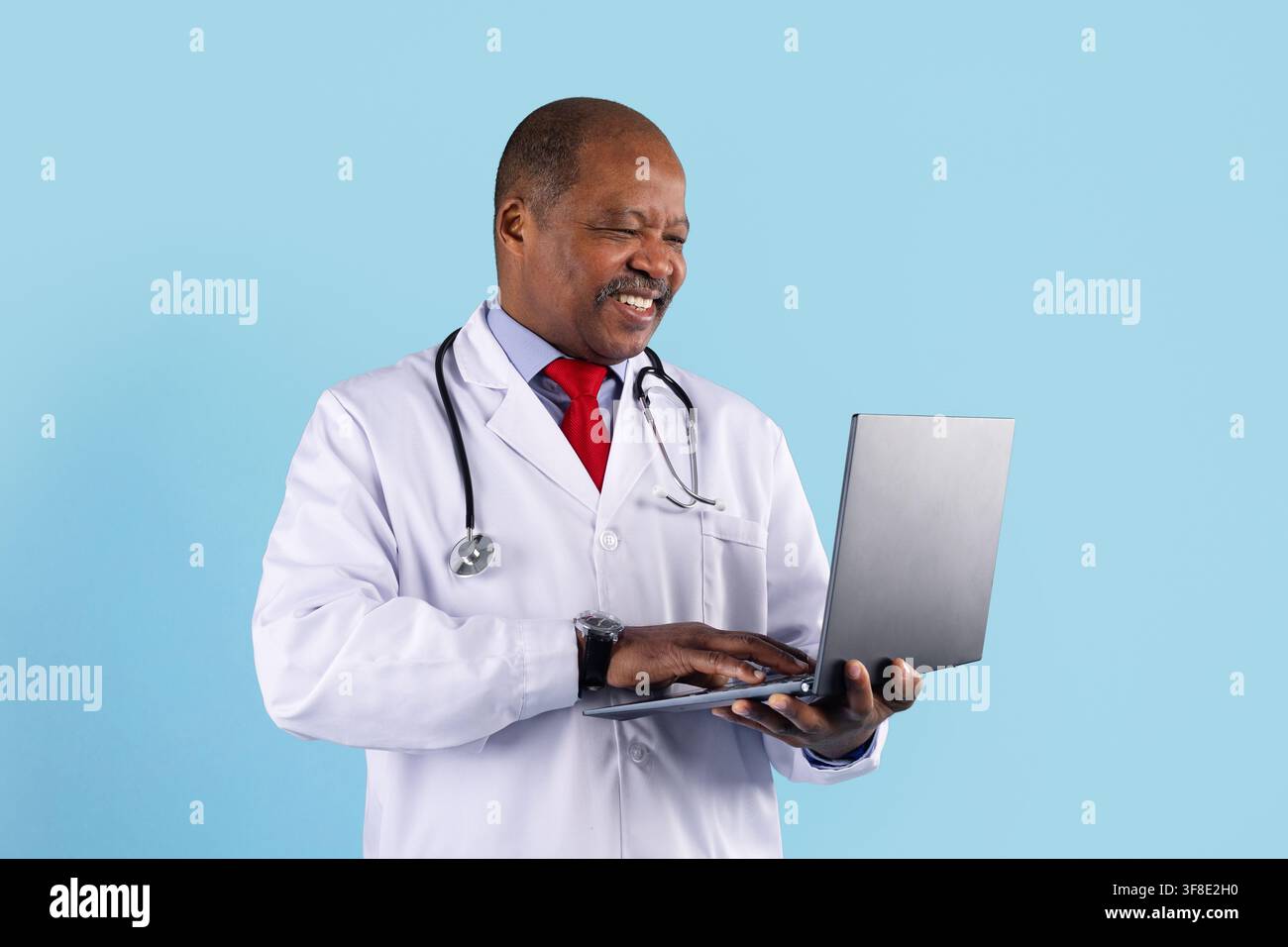 Smiling mature African American doctor using laptop on blue studio background Stock Photo