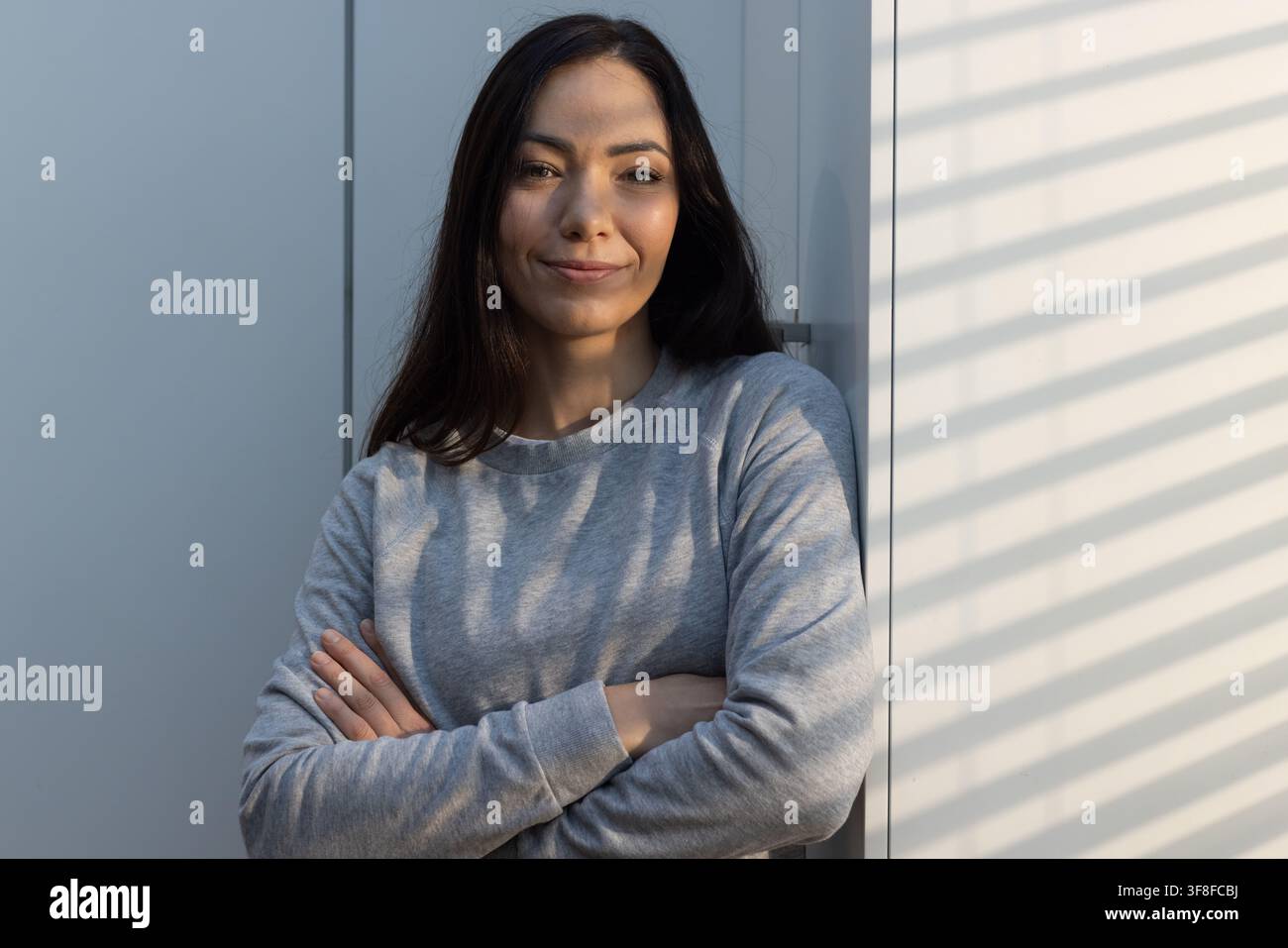 Woman leaning against light-colored panel near window, blinds casting striped shadows on sweatshirt Stock Photo