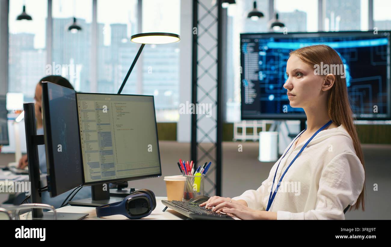 Female technician coding with AI language model assistance in startup office, using PC. Intern using computer, integrating artificial intelligence LLM assistant suggestions into code editor, camera A Stock Photo