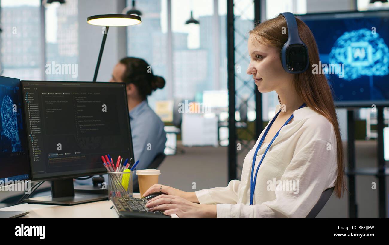 Young female programmer in office reviewing AI system metrics, listening music, monitoring performance dashboards. Junior dev wearing headphones tuning neural networks using tools on PC, camera A Stock Photo