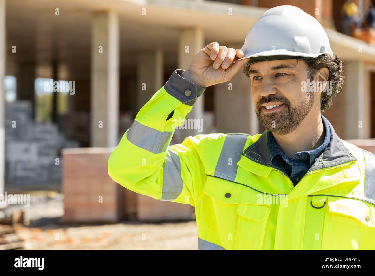 Bearded construction worker touching hardhat and looking away Stock Photo