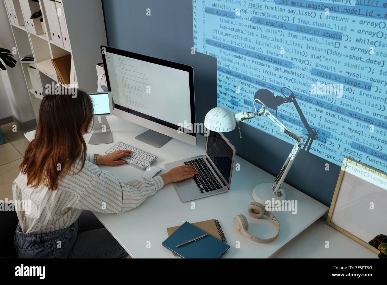 Female software developer coding on multiple screens with projected code in a modern office environment, Lines of code are projected onto the wall, th Stock Photo