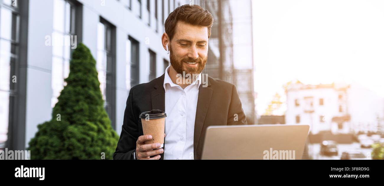 Happy Caucasian Man Using Laptop And Drinking Coffee Outdoors In Modern Business District Stock Photo