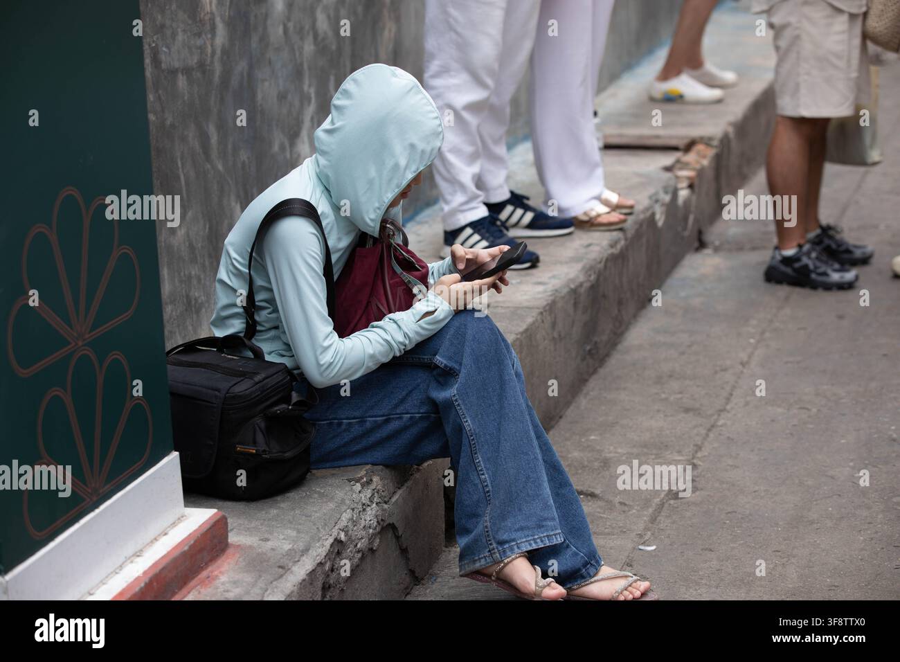 Woman using smartphone while sitting on sidewalk, side profile. Stock Photo
