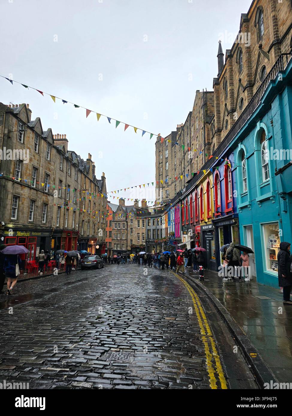 Rain-soaked cobblestones reflect the light on Victoria Street, lined with brightly painted shopfronts and strings of bunting in the Old Town, Edinburg - Smartphone Captured Stock Image