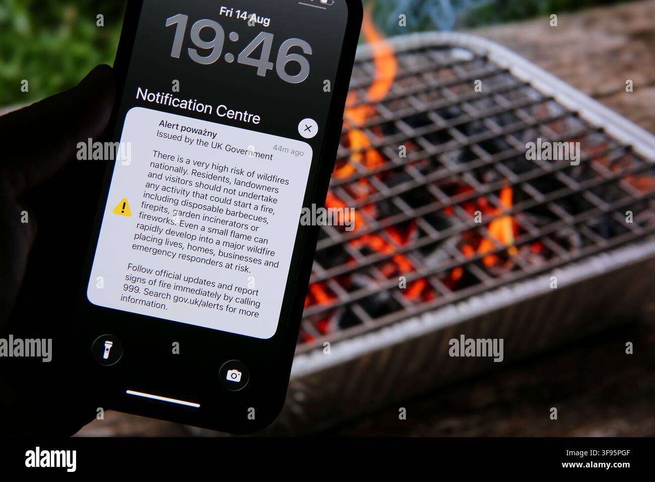 London, UK. 14 August 2026. A mobile phone displays a UK Government Emergency Alert warning of a very high risk of wildfires, beside a burning disposable barbecue in the UK. The alert was sent across England and Wales as extreme heat and dry conditions fuel a surge in wildfires. The government has also introduced a temporary ban on disposable barbecue sales, while authorities urge the public to avoid activities that could start fires. Credit: Waldemar Sikora / Alamy Live News Stock Photo